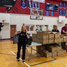 Volunteer with two filled backpacks over her shoulder as she stands in front of a table full of boxes with supplies in them